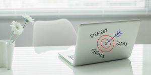 Heller Arbeitsplatz mit Laptop, auf dem eine gezeichnete Zielscheibe mit Pfeil sowie die Begriffe „Strategy“, „Plans“ und „Goals“ zu sehen sind.